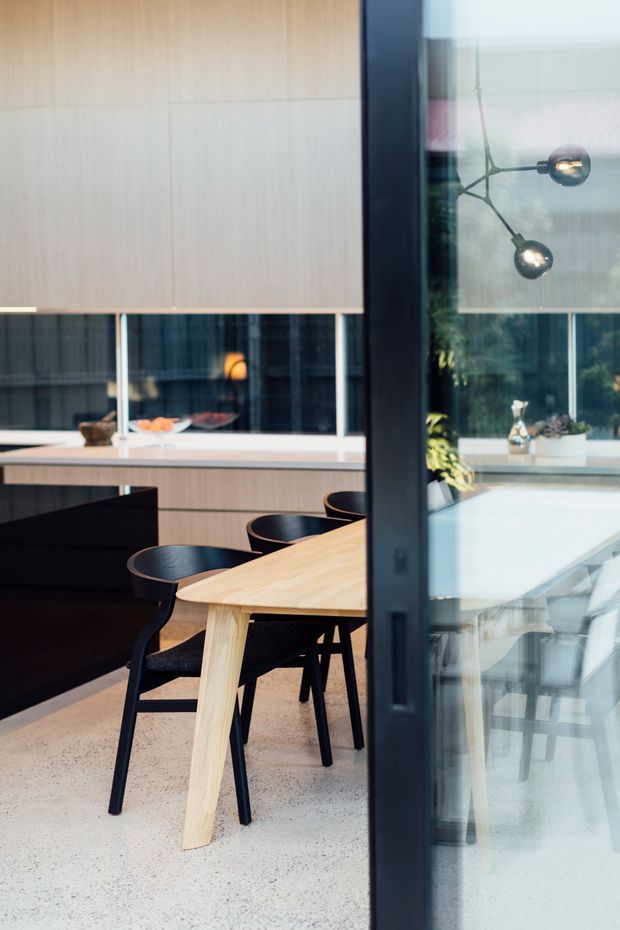 Contemporary dining area viewed through a dark-framed glazed partition or doorway. The room features a large light timber dining table with black dining chairs, a dark kitchen island, pale timber cabinetry, expansive internal or external glazing, and a sculptural black multi-arm pendant light. The floor appears to be a pale polished or smooth concrete/terrazzo-like finish, and reflected glazing adds depth to the scene.