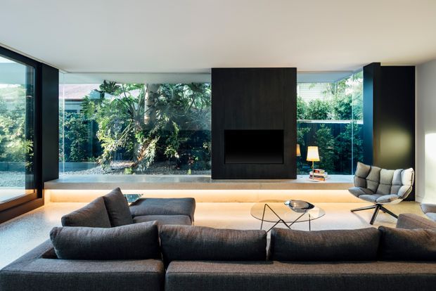 Contemporary living room with a large dark sectional sofa arranged around a low glass coffee table, a wall-mounted fireplace set within a dark feature panel, and expansive floor-to-ceiling glazing overlooking a landscaped garden. Warm concealed lighting runs beneath the fireplace wall, while a sculptural lounge chair and side table occupy the right side of the room.