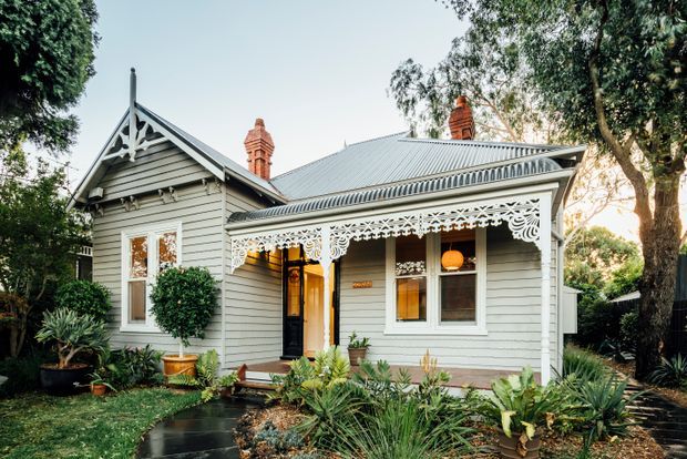 Exterior view of a character weatherboard cottage surrounded by mature garden planting. The home features a steep gabled roof, corrugated metal roofing, ornate white timber fretwork along the verandah, decorative gable detailing, brick chimneys, white-trimmed windows, and a pale grey painted facade.