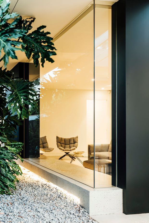 Exterior view through a large floor-to-ceiling glazed wall into a refined living room. The interior features warm cream walls and ceiling, dark-framed glazing, a pale floor, sculptural tufted lounge chairs, a low glass coffee table, and a sofa. Dense leafy planting borders the facade, with a light gravel bed and a raised pale concrete plinth outside. The image emphasises the relationship between the landscaped exterior and the bright, minimalist interior.