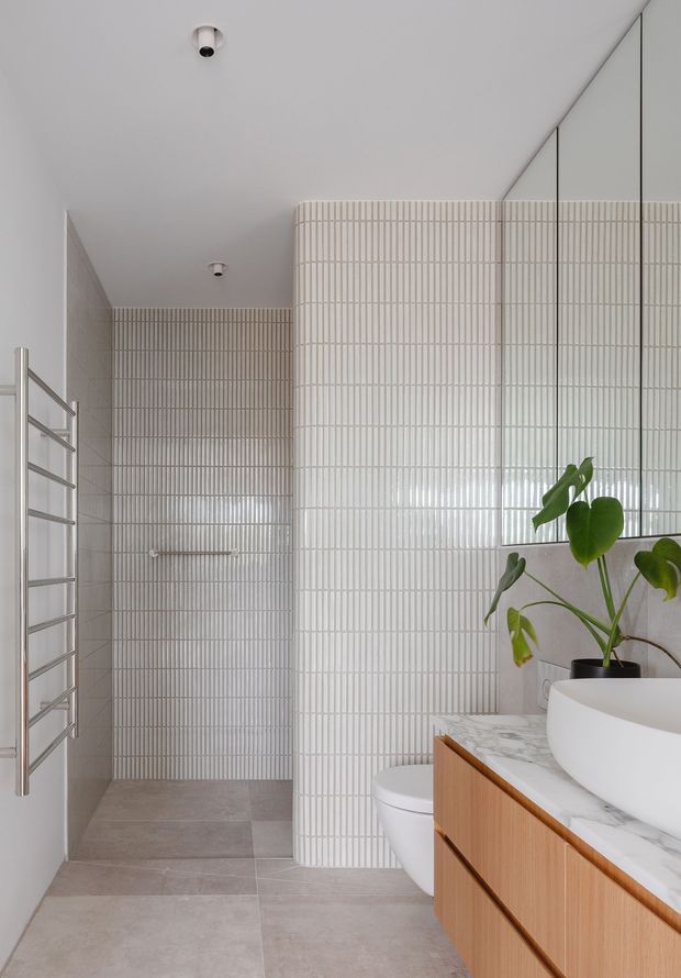 Bright contemporary bathroom featuring a walk-in shower enclosed by curved, vertically ribbed white tiles, a frameless glass partition, stainless steel heated towel rail, pale stone-look floor tiles, and a floating timber vanity with a white vessel basin and marble-look benchtop. A potted plant adds greenery.