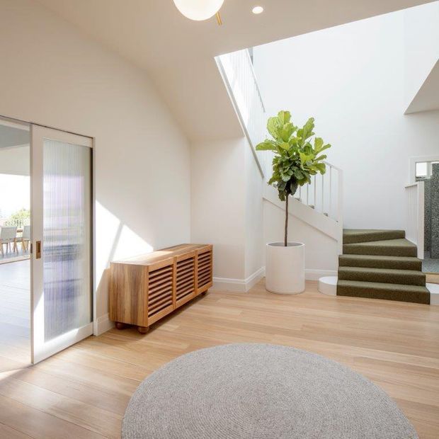 Bright contemporary entryway with pale timber flooring, white walls and a sculptural open stair. A slatted timber sideboard sits beside a frosted-glass sliding door, while a large potted fiddle-leaf fig adds greenery. The stair is carpeted in a muted olive-grey tone and a round pale rug anchors the foreground.