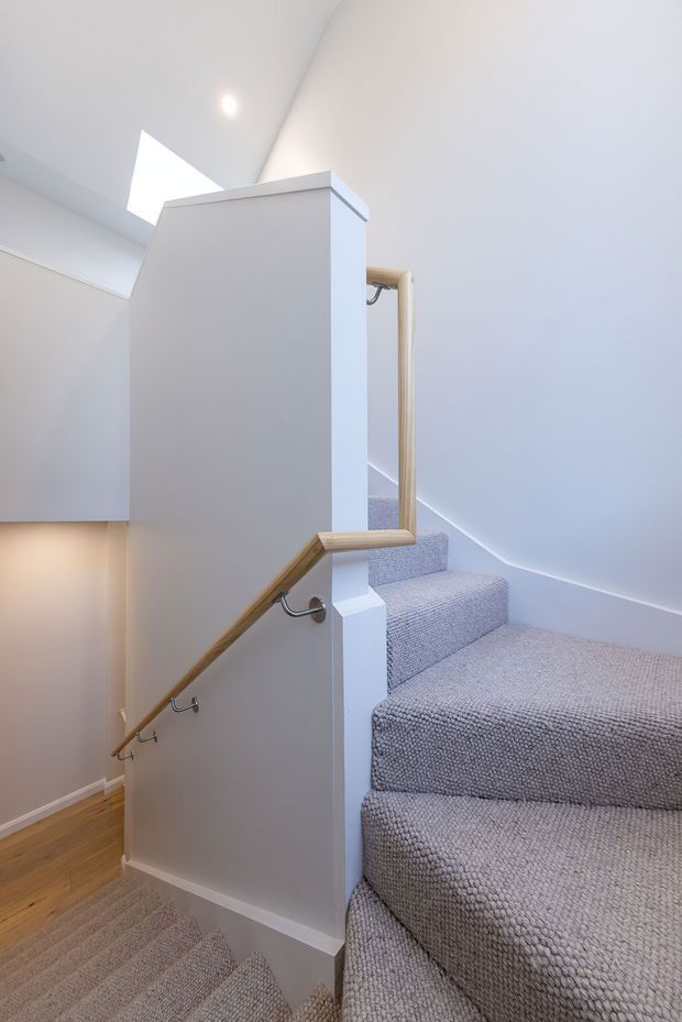 A bright, narrow residential stairway with carpeted steps and landings, white painted walls, and a central half-height partition. Timber handrails with metal brackets run along both sides, while a skylight brings natural light into the stairwell. Timber flooring is visible at the lower level.