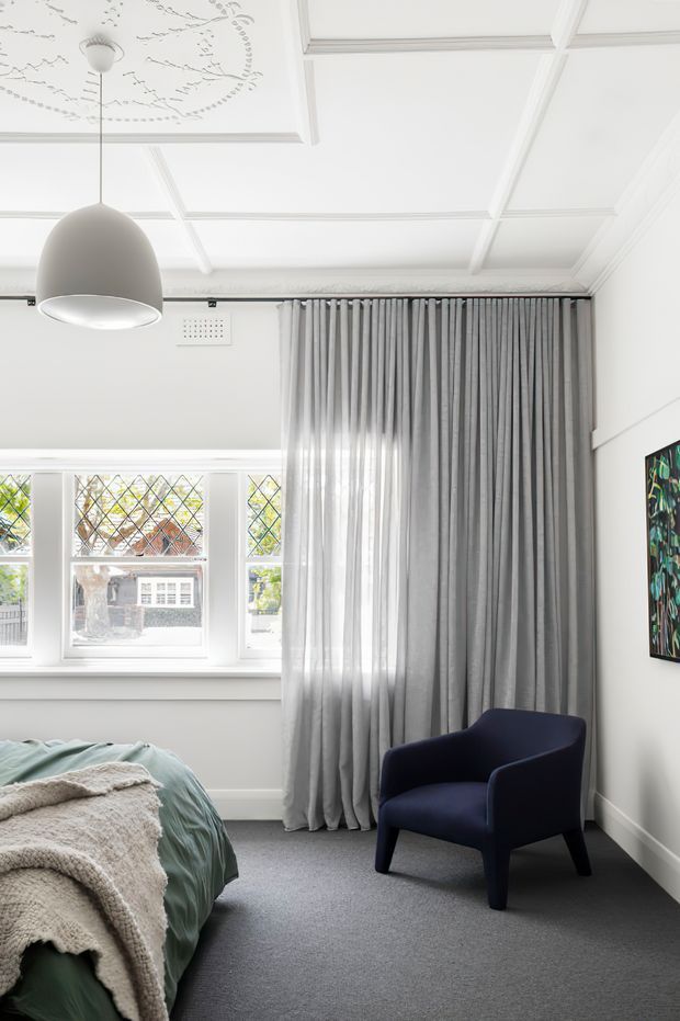 Bright, refined bedroom with a grey carpet, pale walls, decorative white coffered ceiling, tall grey curtains, and leadlight-style windows. A partially visible bed is dressed in muted green and cream bedding, complemented by a navy upholstered armchair and a colourful wall artwork.