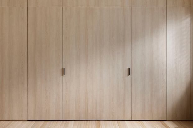 A close-up of a minimalist built-in timber wardrobe or storage wall with full-height, light natural-wood cabinet doors and slim dark vertical handles. Matching light timber flooring is visible at the base. The composition is clean and understated, with soft natural lighting and minimal detailing.