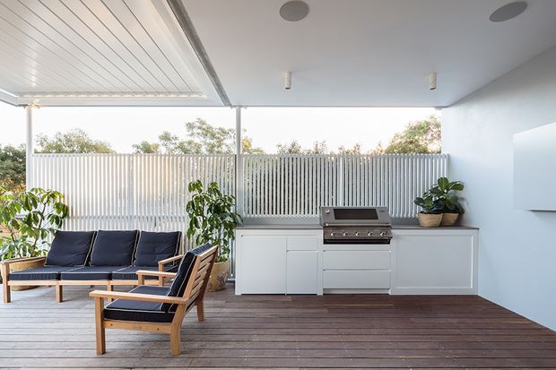 Covered outdoor entertaining area with a built-in barbecue kitchen, white cabinetry, timber decking, slatted privacy screening, and a mid-century-inspired outdoor lounge setting. The space is bright and open, with potted plants and a clean contemporary finish.