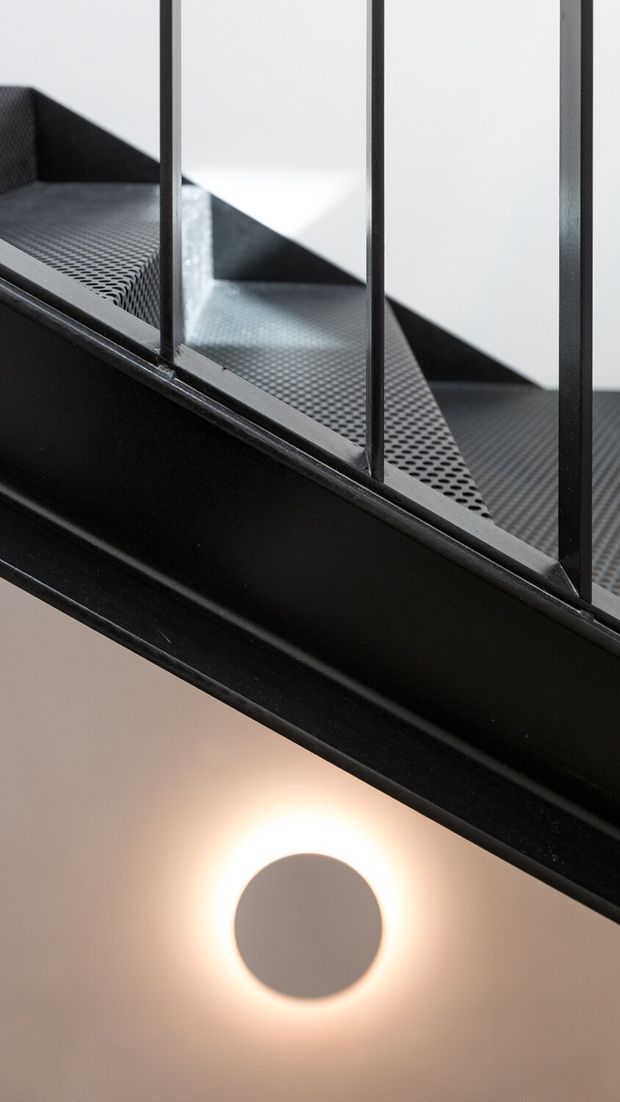 Close-up architectural detail of a black metal stair or mezzanine structure with perforated metal treads, solid steel stringers, and simple vertical balustrade rails. A round wall or ceiling light glows beneath the structure, creating a stark industrial composition against pale surfaces.