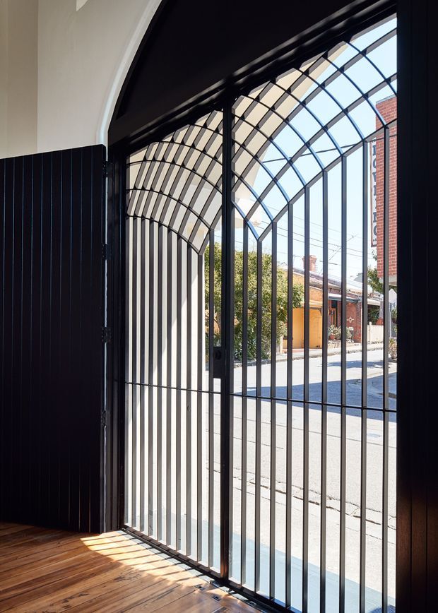 Interior view toward a tall black metal security gate with an arched, curved-grid upper section. The gate opens onto a bright residential street, with brick homes visible outside. Inside, the entry has warm timber flooring, white rendered walls, and dark vertical timber or metal wall panelling. Strong sunlight casts the gate's linear shadows across the floor.
