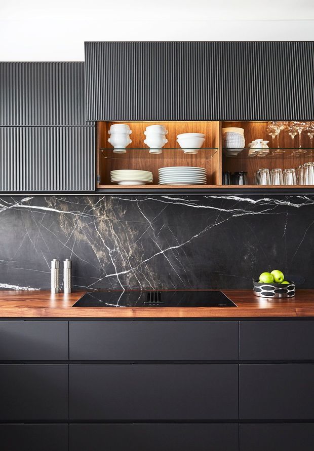 A refined contemporary kitchen featuring matte dark cabinetry, a rich timber benchtop, and a dramatic black marble-look splashback with white veining. Vertically ribbed dark wall cabinetry and open timber-lined display shelving showcase white crockery and glassware. A black induction cooktop, chrome utensils, and a bowl of green fruit complete the composition.