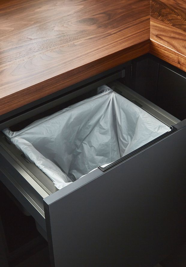 Close-up of a built-in pull-out waste bin drawer with a fitted liner, set beneath a thick dark timber benchtop. The drawer fronts and surrounding cabinetry are matte dark charcoal, with brushed metal drawer hardware and trim. The image focuses on kitchen joinery functionality.