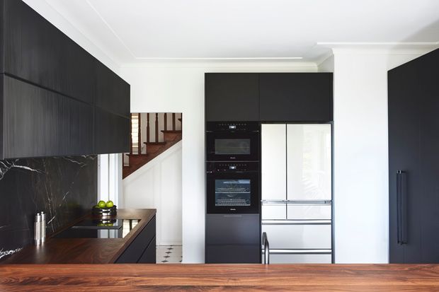 Contemporary kitchen with dark timber cabinetry, a dramatic black marble-look splashback, integrated black appliances, and a large stainless-steel refrigerator. A warm timber island or benchtop occupies the foreground, while a staircase is visible beyond the kitchen.