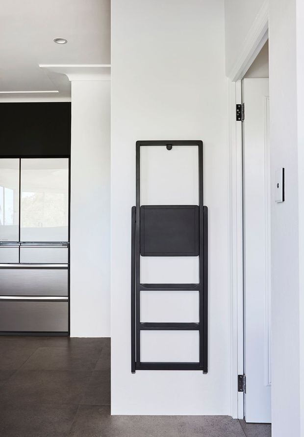 A minimalist interior hallway or entry area with white walls, dark tiled flooring, and a black wall-mounted folding drying rack or utility rail. An open white door is visible on the right, while a dark-framed glazed opening and built-in cabinetry appear to the left.