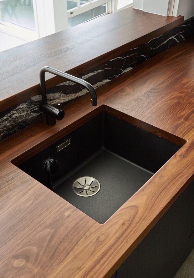 Close-up view of a refined kitchen work area featuring a large black undermount sink, matte black faucet and fixtures, warm timber benchtops, dark cabinetry, and a striking black marble splashback with white veining. Natural light enters through the adjacent windows.