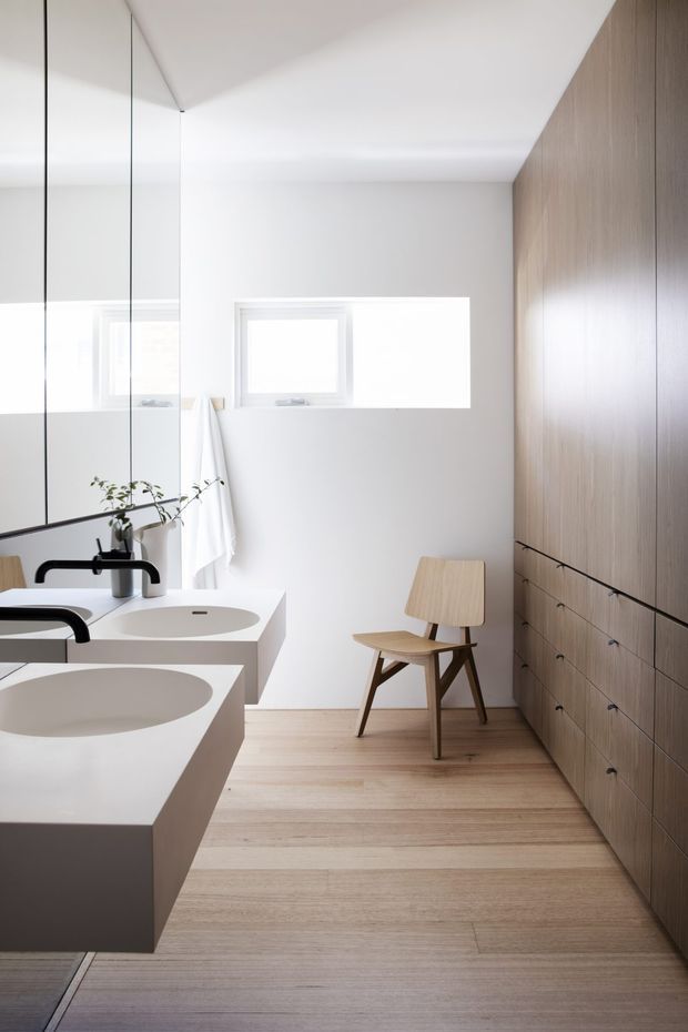 Bright minimalist bathroom with twin white integrated vanity basins and matte black wall-mounted tapware in the foreground. A freestanding white bathtub is partially visible behind, with a small high window, white walls, a full-height mirrored surface, pale timber flooring, and extensive warm timber built-in cabinetry with drawers along the right wall. A simple timber chair and towel add functional detail.