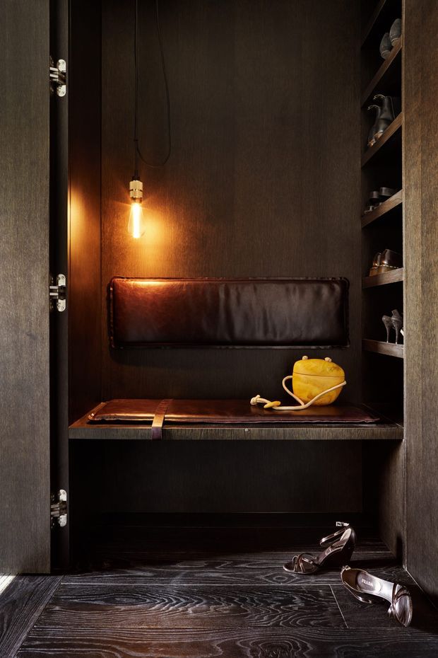 A dark, compact built-in wardrobe or dressing nook with deep timber-toned cabinetry, open shoe shelves, a leather-upholstered bench, and a warm exposed pendant light. A yellow handbag and shoes sit on the bench and floor, creating a moody boutique-style dressing area.