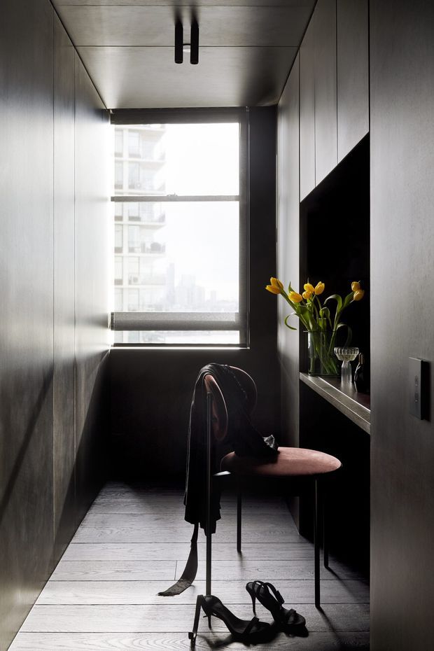 A narrow, moody interior nook or hallway with dark vertically panelled walls, a pale timber-look floor, and a large black-framed window overlooking a high-rise urban setting. A slim console shelf supports a wall-mounted black screen, vase of yellow flowers, and small objects. A burgundy upholstered chair with clothing draped over it and black high-heeled shoes on the floor create a styled dressing or entry area. Minimal black ceiling lighting and strong daylight contrast define the composition.