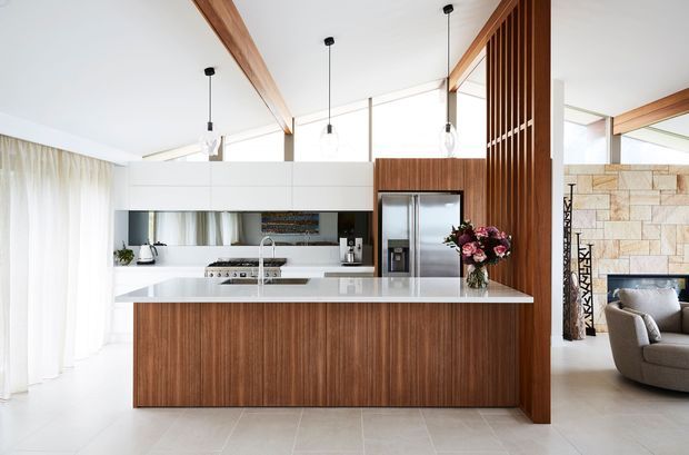 Bright open-plan kitchen with a large white island, warm timber veneer cabinetry, integrated stainless-steel appliances, white overhead joinery, pendant lighting and timber screening. A living area with a fireplace and stone feature wall is visible beyond the kitchen.
