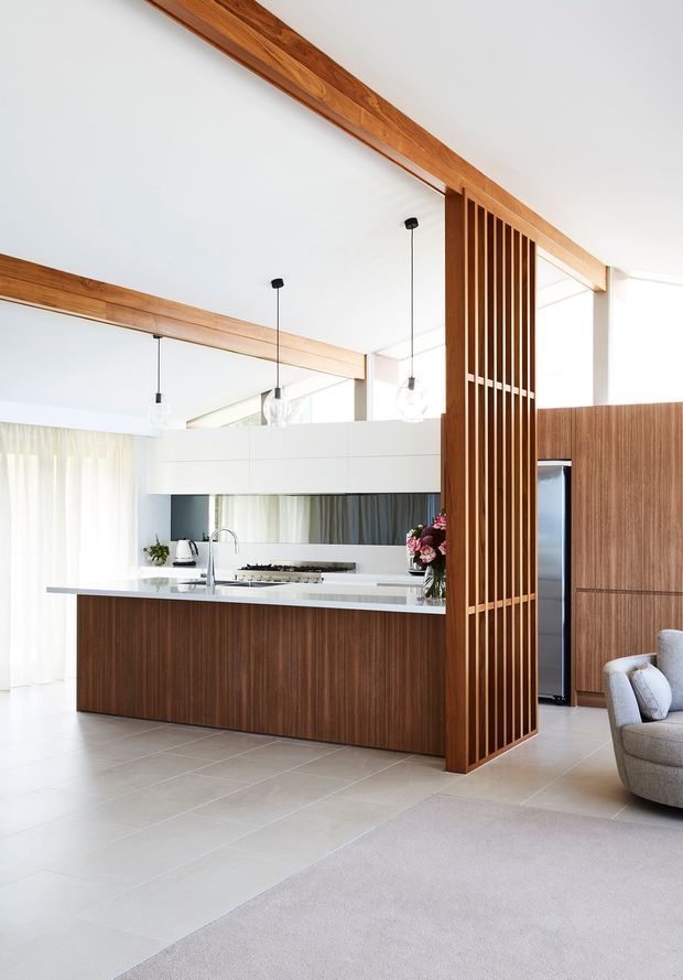 Bright open-plan kitchen and living area featuring a large dark timber island with a white stone benchtop, integrated cooking and sink zones, full-height timber cabinetry, and a vertical timber slatted room divider. White walls and ceiling, exposed timber beams, pendant lights, pale tiled flooring, and soft neutral furnishings create a refined contemporary interior.
