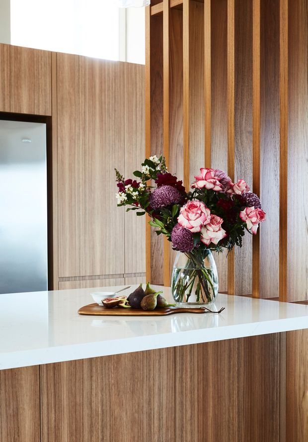 A bright, refined kitchen interior featuring warm vertical timber cabinetry and a matching slatted timber feature wall. A glossy white island benchtop dominates the foreground, styled with a glass vase of flowers and a wooden serving board with fruit. A tall dark-framed opening or appliance enclosure is visible at left.