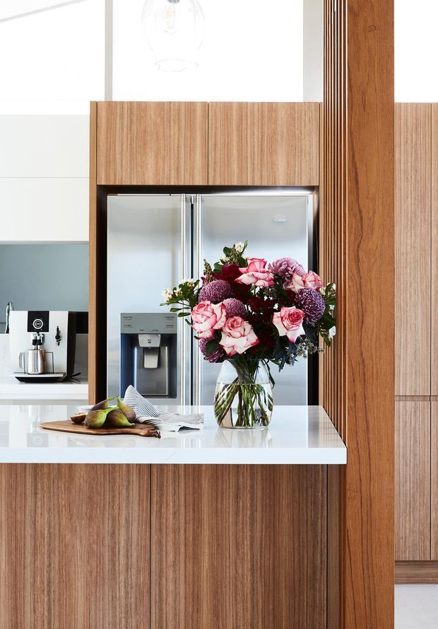 A bright contemporary kitchen with extensive vertical-grain timber cabinetry, a white stone island benchtop, integrated stainless-steel double-door refrigerator, and a coffee station set against a muted blue-green backsplash. A floral arrangement and fruit sit on the island, while a timber screen or structural post adds vertical definition.