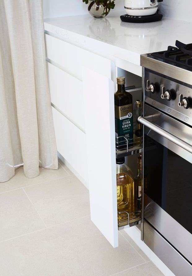Close-up of a bright white kitchen featuring a glossy white stone benchtop, handleless white cabinetry, pale tiled flooring, and a stainless-steel built-in gas oven. A narrow pull-out pantry beside the oven is open, showing oil bottles and wire storage racks. Sheer curtains and small countertop appliances are visible.