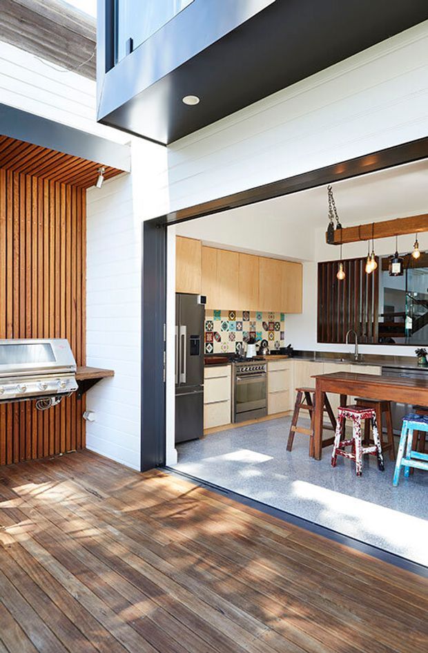 Bright architectural photograph showing a contemporary indoor-outdoor entertaining area. A timber-clad patio or deck with built-in stainless-steel barbecue opens directly into a kitchen through a large dark-framed opening. The kitchen includes light timber cabinetry, a patterned tile splashback, stainless-steel appliances, a central island with seating, pendant lights, and dark vertical timber screening. A double-height void and upper-level glazing emphasize the open plan.