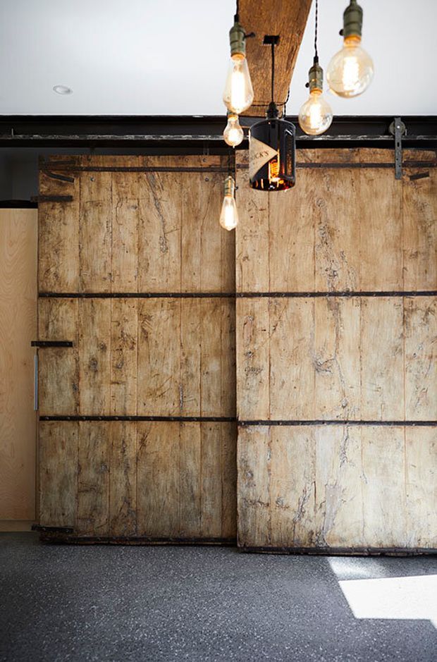 Interior view dominated by large rustic sliding barn doors made from weathered timber, set within a dark industrial-style track and frame. Several exposed Edison-style pendant bulbs hang from a substantial timber beam, while the floor is dark and speckled. The composition highlights reclaimed materials, visible hardware, and an industrial-rustic aesthetic.