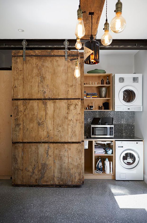 A compact industrial-style laundry with stacked white washing and drying appliances, open timber shelving, a microwave, and a large weathered timber barn door on exposed metal sliding hardware. The room features dark patterned tile backsplash, a grey speckled floor, exposed timber and steel elements, and pendant lighting made from hanging filament bulbs and repurposed bottles.