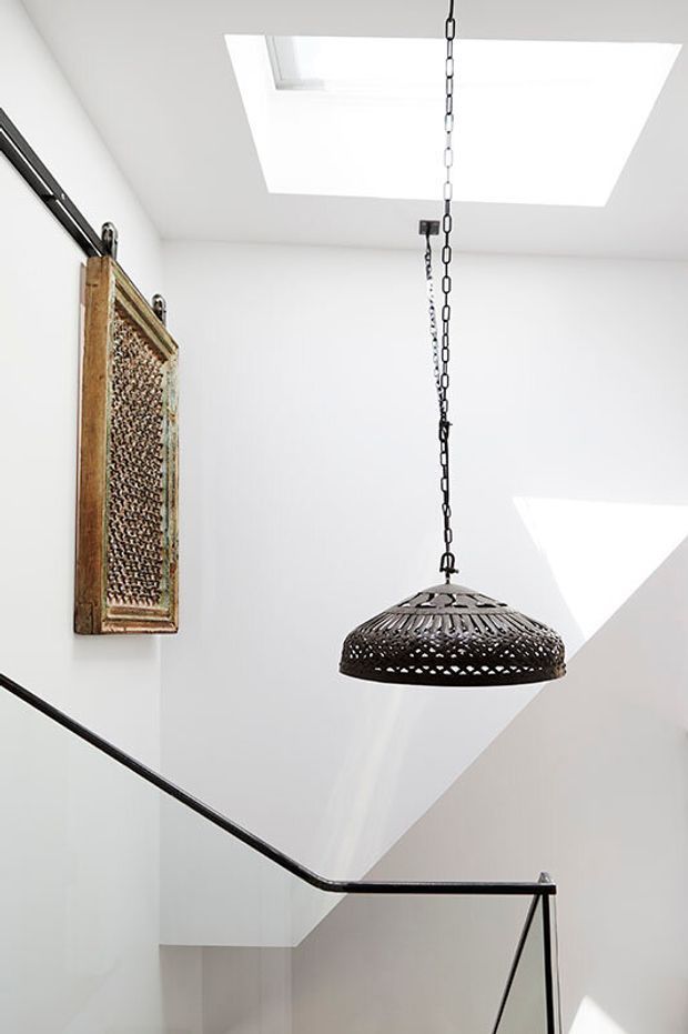 Bright minimalist stairwell with white walls and geometric architectural forms, featuring a large rectangular skylight, angular wall openings, black metal handrails, and a suspended woven pendant light. A decorative carved wall panel adds a rustic, artisanal accent.