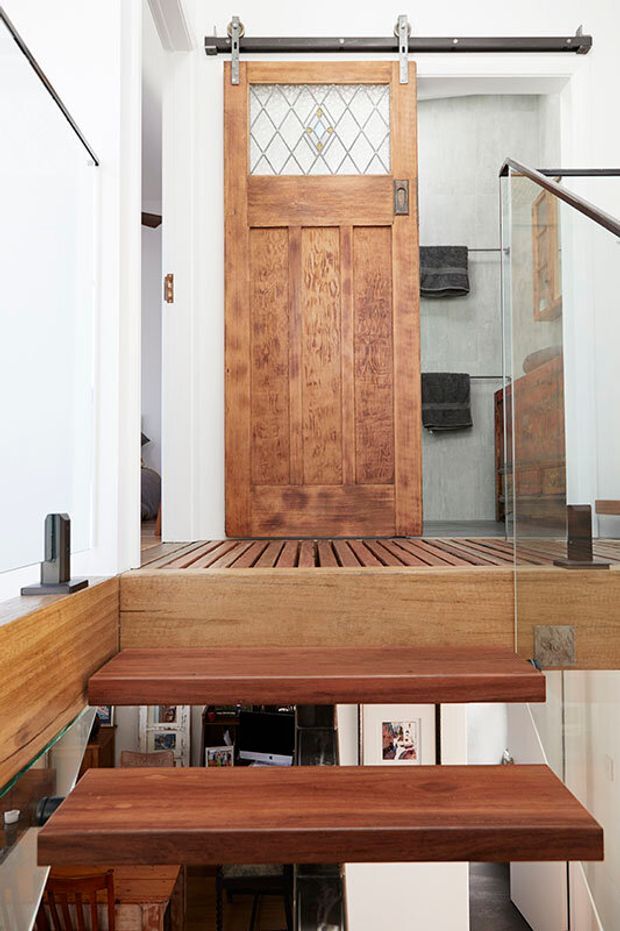 Interior view of a compact stairway with floating timber treads, glass balustrades, and a timber-framed sliding barn door with decorative leaded-style glazing. The stairs connect to an upper-level bathroom, with white walls and warm wood finishes throughout.