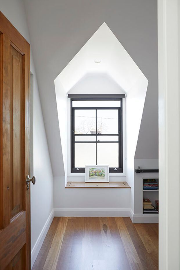 Bright, narrow interior nook or hallway with a distinctive peaked, chapel-like window reveal. The space has warm timber flooring, a painted white built-in window seat or sill, dark-framed divided glazing, a timber door at left, and a small recessed bookshelf at right. The high angular ceiling and crisp white walls create a clean architectural focal point.