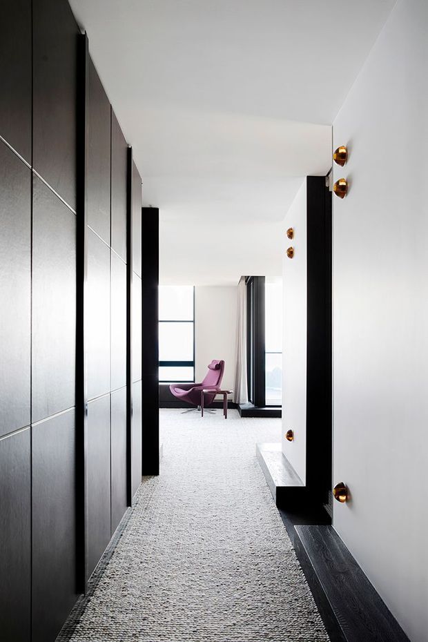 A long, narrow contemporary hallway with floor-to-ceiling dark built-in wall panelling on the left, white walls and ceiling, dark timber flooring at the edges, and a textured light runner. Black-framed glazing and curtains open toward a bright sitting area at the end, furnished with a distinctive magenta lounge chair. Small brass wall lights provide warm accents along the right wall.