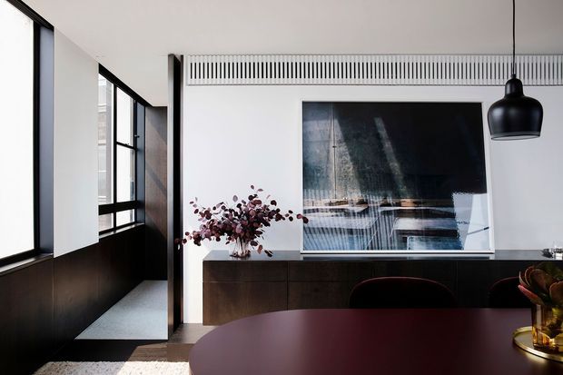 A refined contemporary dining area with a dark round dining table in the foreground, a low dark timber sideboard, sculptural black pendant light, large abstract artwork, and floor-to-ceiling black-framed glazing along the left side. A narrow adjacent walkway or entry passage is visible beyond the dining space. The palette is predominantly white, black, and dark timber, with carefully integrated linear ventilation detailing above the artwork.