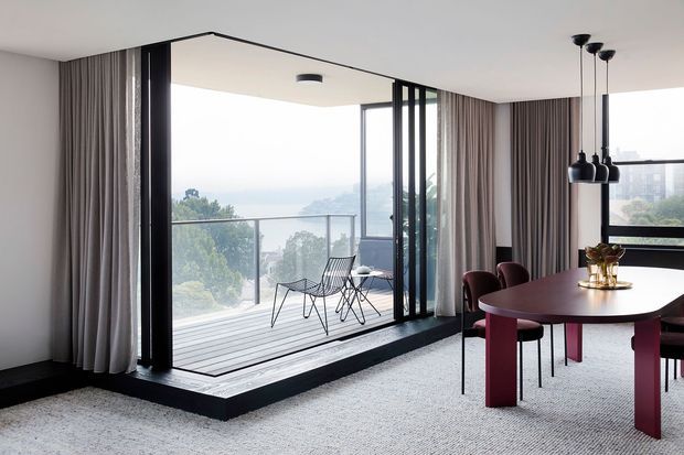 A refined contemporary dining area with a large corner opening to a glazed balcony or terrace. Floor-to-ceiling black-framed glazing and slim vertical black screens frame expansive views, while full-height taupe curtains, pale carpet, and a round dark dining table with burgundy chairs create a polished, softly monochromatic interior. Pendant lights hang above the dining table, and outdoor seating is visible beyond the glazing.