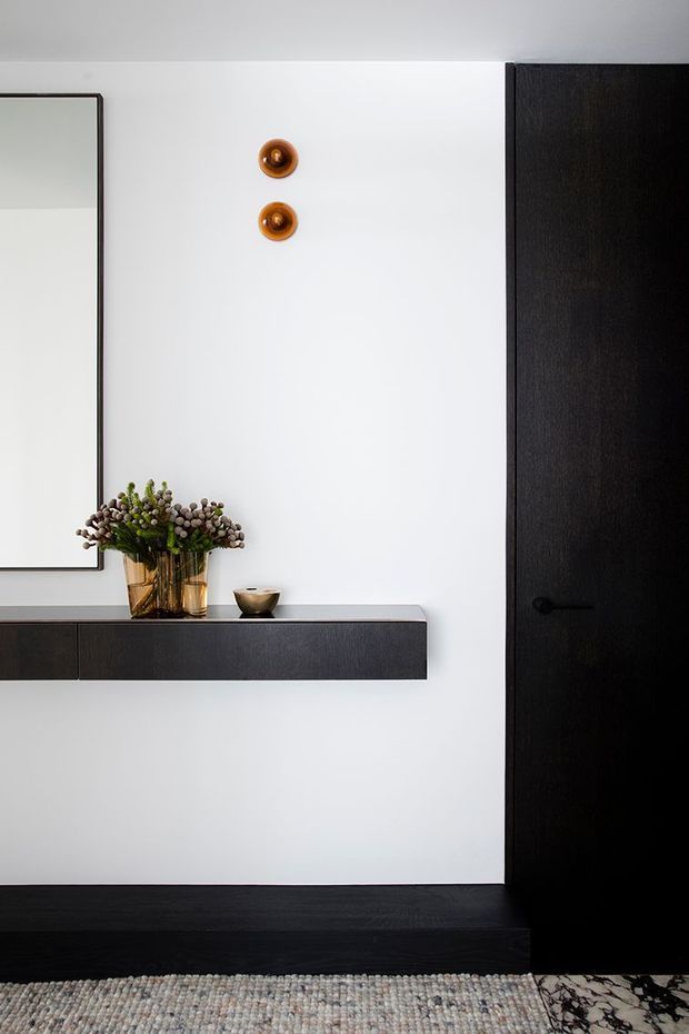 A minimalist entryway or hallway with a crisp white wall, a large black-framed mirror, and a floating dark timber console. A black timber door and dark skirting create strong contrast, while two small brass-toned wall lights, a floral arrangement, and a textured rug add warmth. Marble-patterned flooring is visible at the lower right.