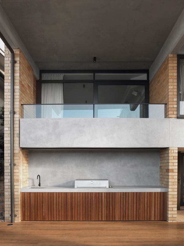 A contemporary multi-level outdoor entertaining area viewed from a covered patio. The lower level features a long built-in outdoor kitchen with vertically slatted timber cabinetry, a pale benchtop, sink and barbecue, set against a smooth grey concrete or rendered wall. Above it is a projecting balcony with a solid pale grey rendered fascia and clear glass balustrade, backed by large dark-framed glazed doors. Warm brick piers and timber decking add contrast, while the covered concrete ceiling creates a sheltered, architectural setting.