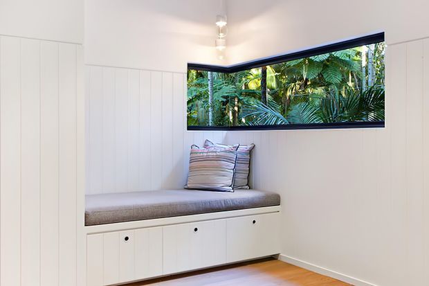 Bright, compact built-in window seat in a white-painted room with vertical wall panelling and integrated storage drawers beneath the upholstered bench. A long horizontal corner window frames lush tropical greenery outside, while a small track light illuminates the clean, minimal space. Timber flooring adds warmth.