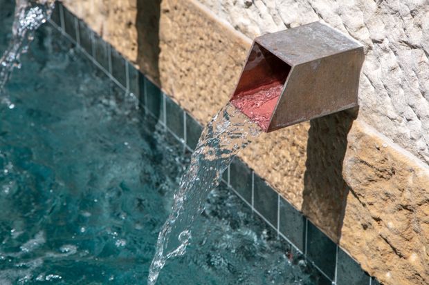 Close-up of a sculptural metal water spout projecting from a rough stone-clad wall, pouring a stream of water into a tiled swimming pool. The image focuses on the pool water feature and contrasting rustic masonry and weathered metal surfaces.
