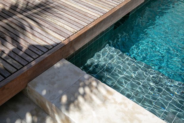 Close-up view of a swimming pool with clear turquoise water, dark green tiled pool walls and floor, and a weathered timber deck with a concrete poolside step or ledge. Dappled shadows fall across the deck and paving, creating a relaxed outdoor setting.
