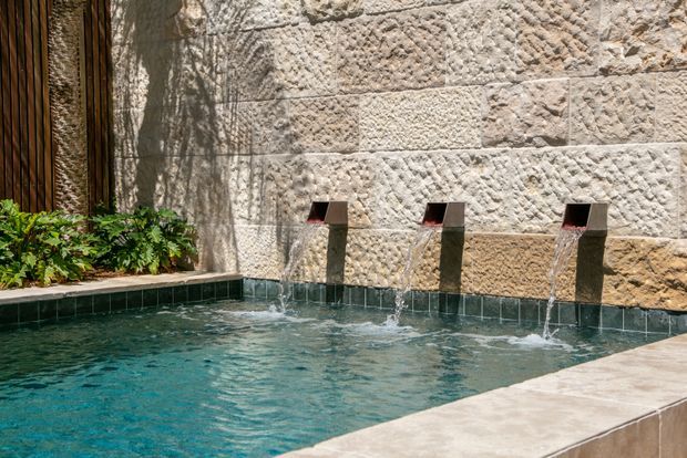 Close-up of a landscaped pool area featuring three rectangular wall-mounted water spouts cascading into a small turquoise pool. The pool is bordered by pale stone coping, a textured rough-cut stone feature wall, dark green tile, timber screening, and dense planting.