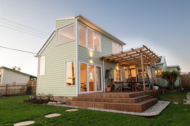 Exterior rear view of a two-storey residential home with pale green horizontal weatherboard cladding, white-trimmed windows and glazed doors, and a timber pergola-covered deck. Broad timber steps lead to a landscaped lawn and gravel-edged garden, with warm interior and exterior lighting creating an inviting evening atmosphere.