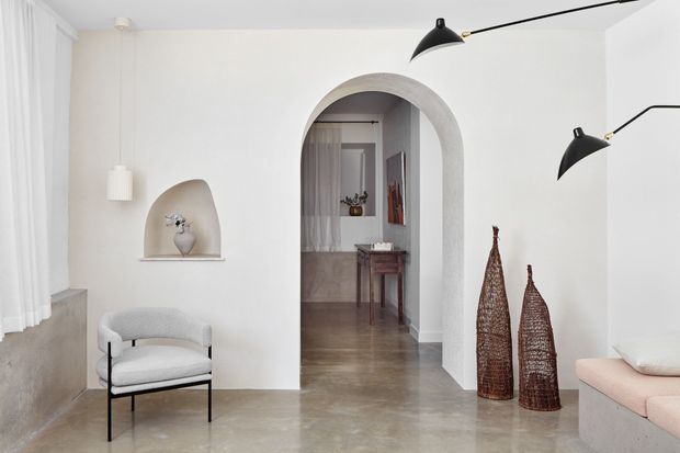 Bright, minimalist interior foyer or living area with white plaster walls, a large rounded archway leading to a secondary space, polished concrete flooring, sheer curtains, sculptural wall niche, black articulated wall lamps, a light upholstered chair, woven decorative vessels, and a pale blush built-in bench. The view is composed around the entry-like sitting area and arched passage.