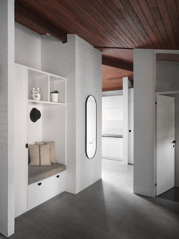 A bright, refined entryway with white-painted brick walls, built-in white storage and a cushioned bench, decorative shelving, an arched mirror, and dark tiled flooring. The space opens toward a minimalist white kitchen or utility area, while a dark timber-lined ceiling adds warmth and contrast.