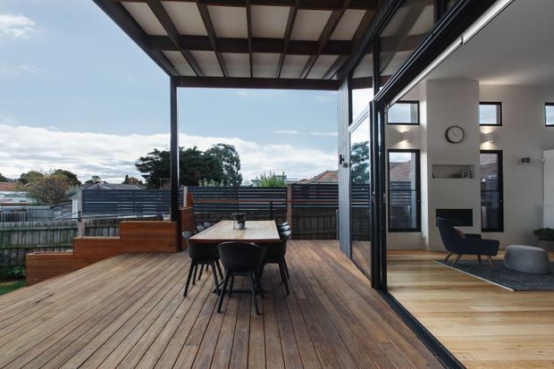 A contemporary indoor-outdoor living space opening onto a large timber deck. The deck includes a dining table and chairs, built-in timber bench seating, and dark horizontal privacy fencing. Floor-to-ceiling glazed sliding doors connect it to a bright interior living area with timber flooring, a fireplace, and modern furnishings. A covered pergola structure extends over the deck.