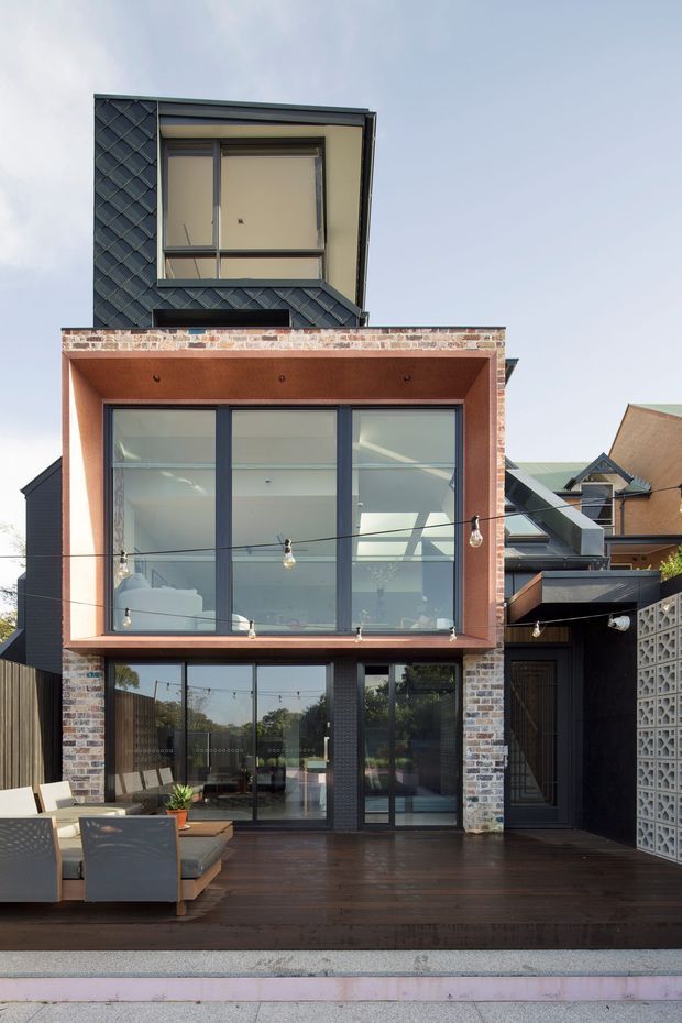 Rear exterior of a narrow multi-storey contemporary dwelling with a projecting upper-level glazed volume, exposed brickwork, dark cladding, and a timber deck furnished with outdoor seating. String lights span the deck, while a patterned concrete-block privacy screen borders the right side.