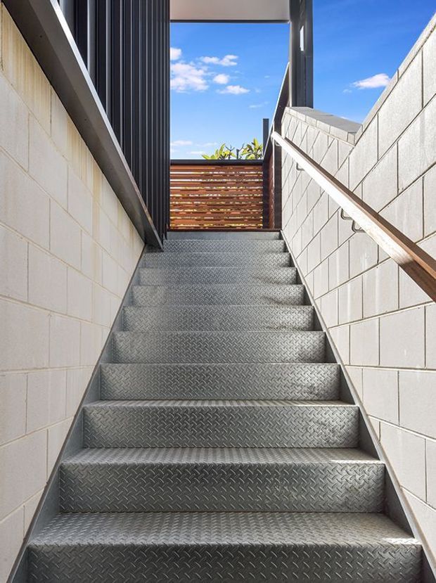 A narrow exterior stairway rises between pale concrete block walls to an upper landing. The stairs have silver diamond-plate metal treads, with a timber handrail on the right wall and dark vertical metal cladding along the left side. A timber slatted screen and blue sky are visible at the top, creating a bright, utilitarian architectural composition.