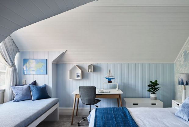 Bright coastal-style bedroom with a built-in window seat, blue and white bedding, a compact desk and task chair, vertical wall panelling, and a sloped paneled ceiling. Nautical decor and soft blue accents create a fresh, relaxed atmosphere.
