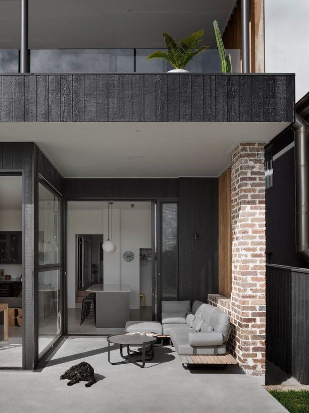 A contemporary covered outdoor living area adjoining a modern brick-and-charred-timber house. The courtyard-style patio has a grey upholstered sofa, nesting coffee tables, glazed sliding doors, dark vertical cladding, a projecting upper-level balcony, exposed brickwork, and potted greenery. The open doors reveal a minimalist kitchen with a central island and pendant light.