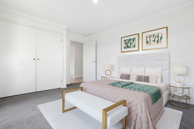 Bright, refined bedroom with a large upholstered bed, pale blush quilted bedding, layered pillows, a white upholstered bench with gold-toned frame, matching bedside tables and lamps, built-in white wardrobes, grey carpet, and framed artwork above the bed. The room has white walls, decorative cornices, and a soft neutral palette with subtle gold accents.