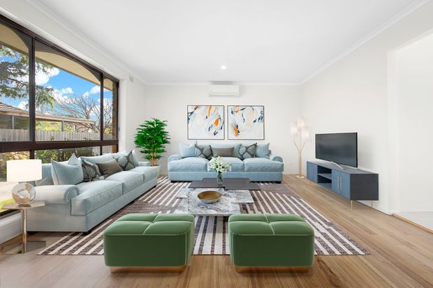 Bright contemporary living room with large dark-framed windows, pale blue sofas, green ottomans, a patterned area rug, timber flooring, abstract artwork, and a compact dark media console. The room has a clean white palette, abundant natural light, and a calm, inviting arrangement.
